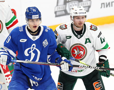 На изображении два хоккеиста. Один в синей форме с номером 19 представляет команду Динамо, другой в белой форме с зелеными элементами и буквой А. Они стоят на льду рядом друг с другом