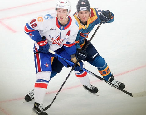 На изображении два хоккеиста в игровой ситуации на льду. Один игрок в бело-синей форме с красными элементами, другой – в синей форме с оранжевыми вставками. Оба держат клюшки, готовясь к игре