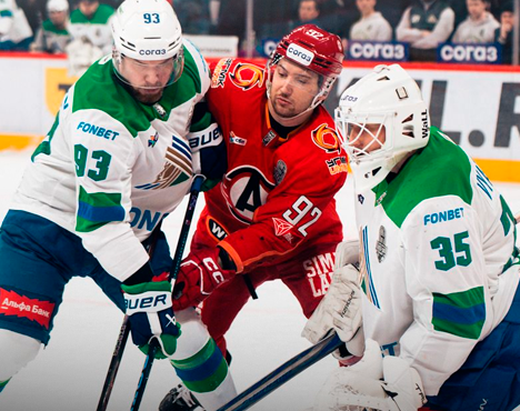 На изображении запечатлен момент хоккейного матча. Игрок в красной форме атакует игроков в бело-зелёной форме, включая вратаря. На формах заметны логотипы команд и спонсоров