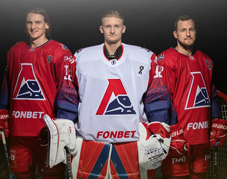 На изображении трое хоккеистов команды Локомотив в спортивной форме: два в красных свитерах и один в белом, вратарь стоит в центре. На форме логотип команды и спонсорская надпись