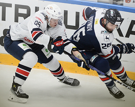 На изображении запечатлен момент хоккейного матча. Два игрока из разных команд в движении: один в белой форме с номером 95, другой в темно-синей форме с номером 26, оба борются за шайбу у борта