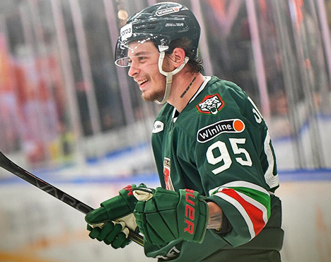 Хоккеист в зелёной форме с номером 95 на спине и шлеме стоит на льду с клюшкой в руке, улыбаясь. На фоне видна хоккейная арена с размытыми деталями