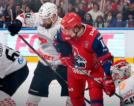 На изображении хоккейный матч. Игроки в форме красного и белого цветов борются за шайбу около ворот. Вратарь команды в белом обмундировании сосредоточенно наблюдает за игрой. Трибуны заполнены зрителями