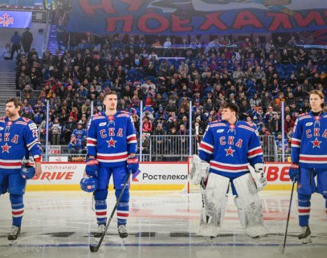 На изображении хоккеисты команды СКА в синей форме с эмблемой на груди стоят на льду перед игрой. На заднем плане — трибуны с болельщиками и баннер с надписью
